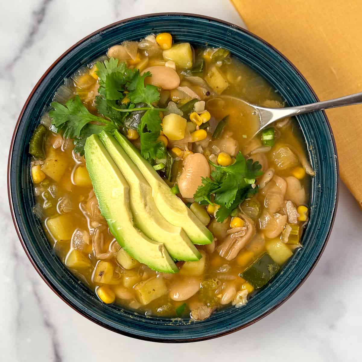 A bowl of vegan white bean chili topped with fresh cilantro and sliced avocado with a spoon on the side.
