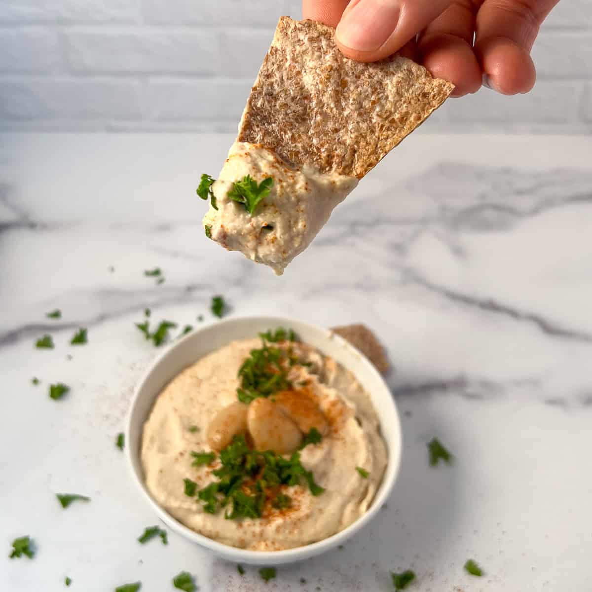 Butter bean hummus in a bowl with a woman's hand dipping a chip in.