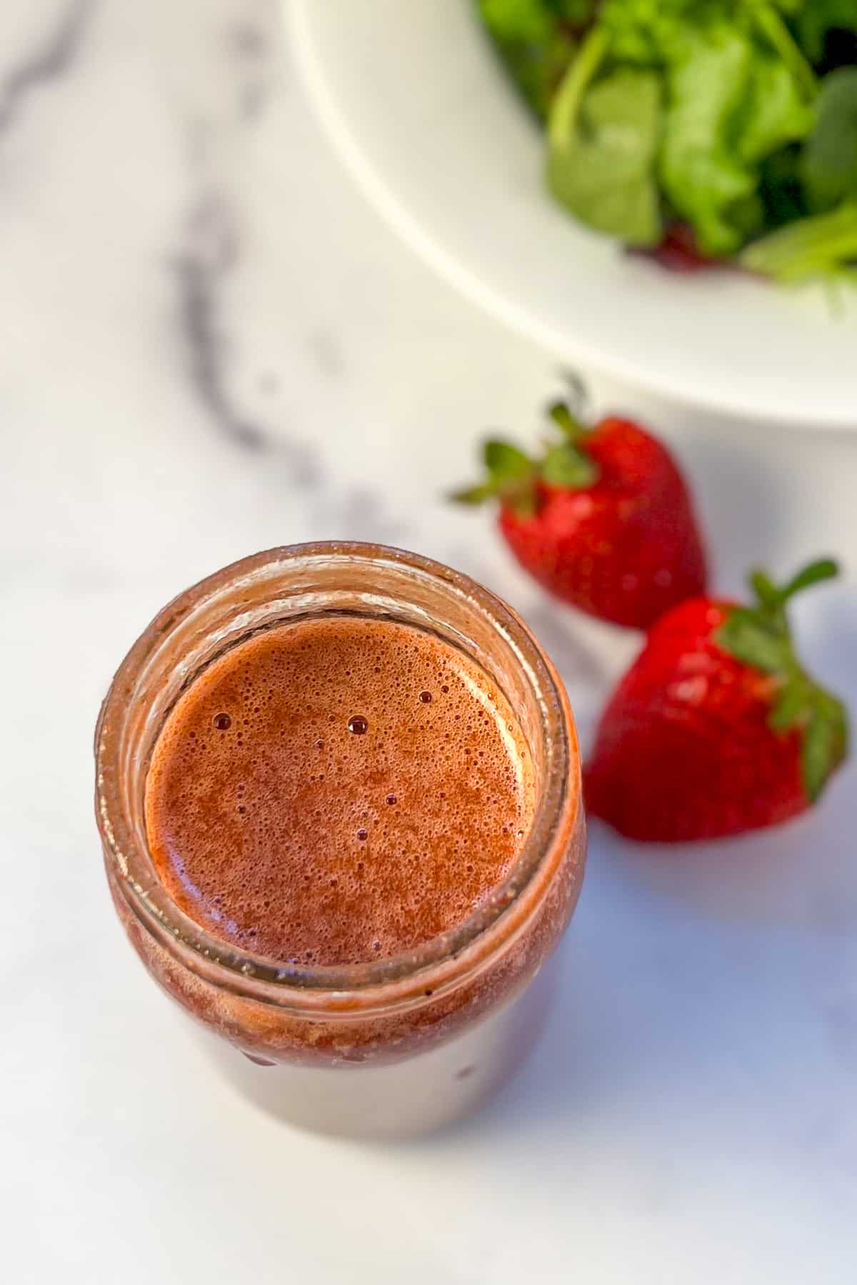 A mason jar with strawberry balsamic salad dressing with fresh strawberries and salad greens on the side.