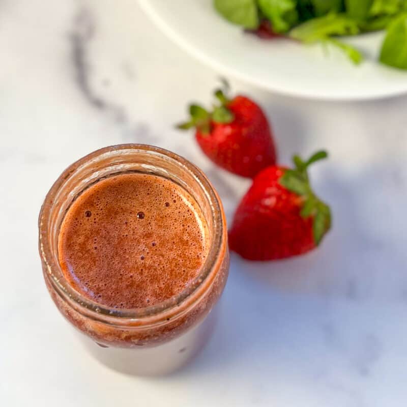 Strawberry balsamic vinegar in a mason jar with fresh strawberries and salad greens blurred in the background.