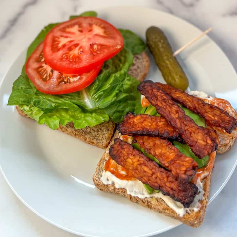 Two slices of whole grain bread; one with lettuce and tomato and the other with sauce and tempeh bacon strips.