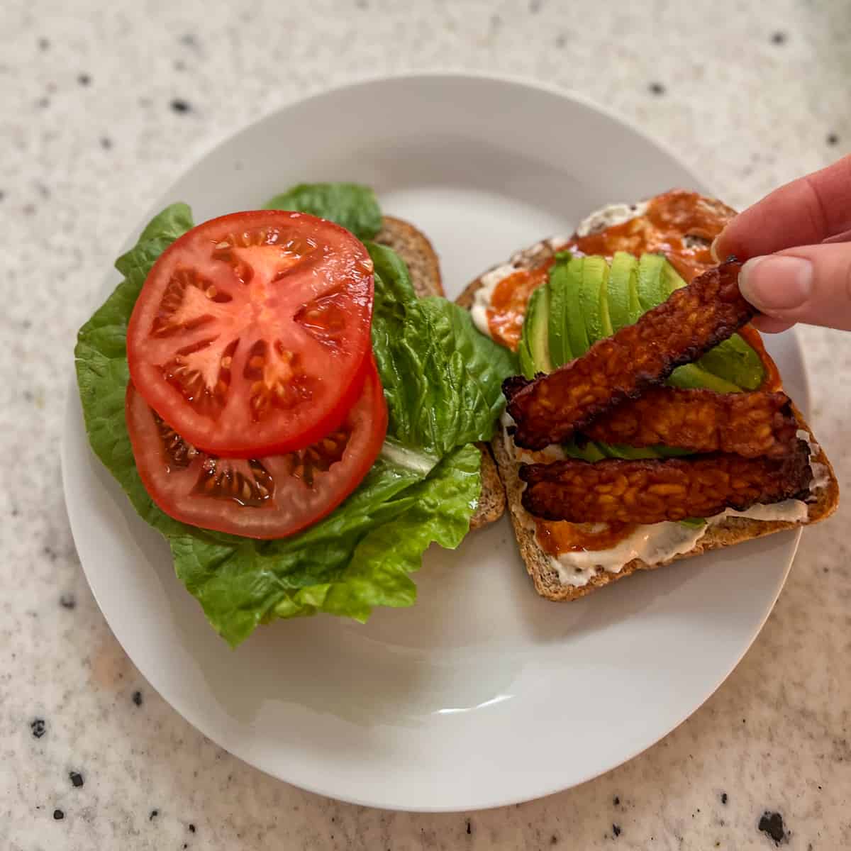 A woman's hand putting a slice of tempeh bacon on the sandwich.