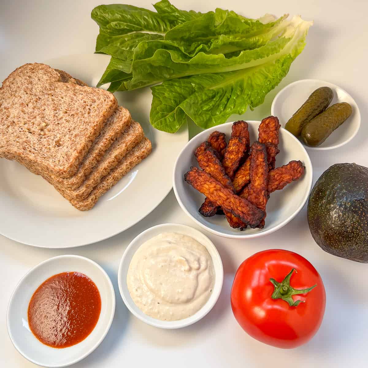 Ingredients for tempeh blt: sprouted grain bread, romaine lettuce, tempeh bacon, vegan mayo, tomato, avocado, dill pickle and sriracha hot sauce.