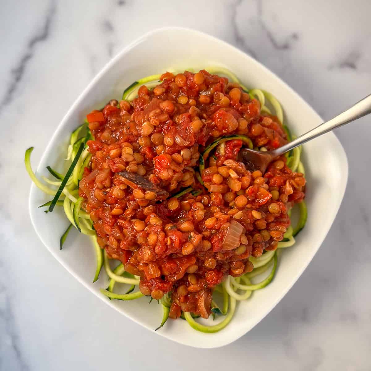 Green lentil Bolognese over raw zucchini pasta in a bowl with fork.