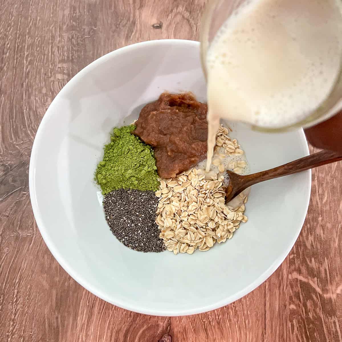 Plant-based milk being poured into a bowl with rolled oats, matcha powder, date syrup and chia seeds.