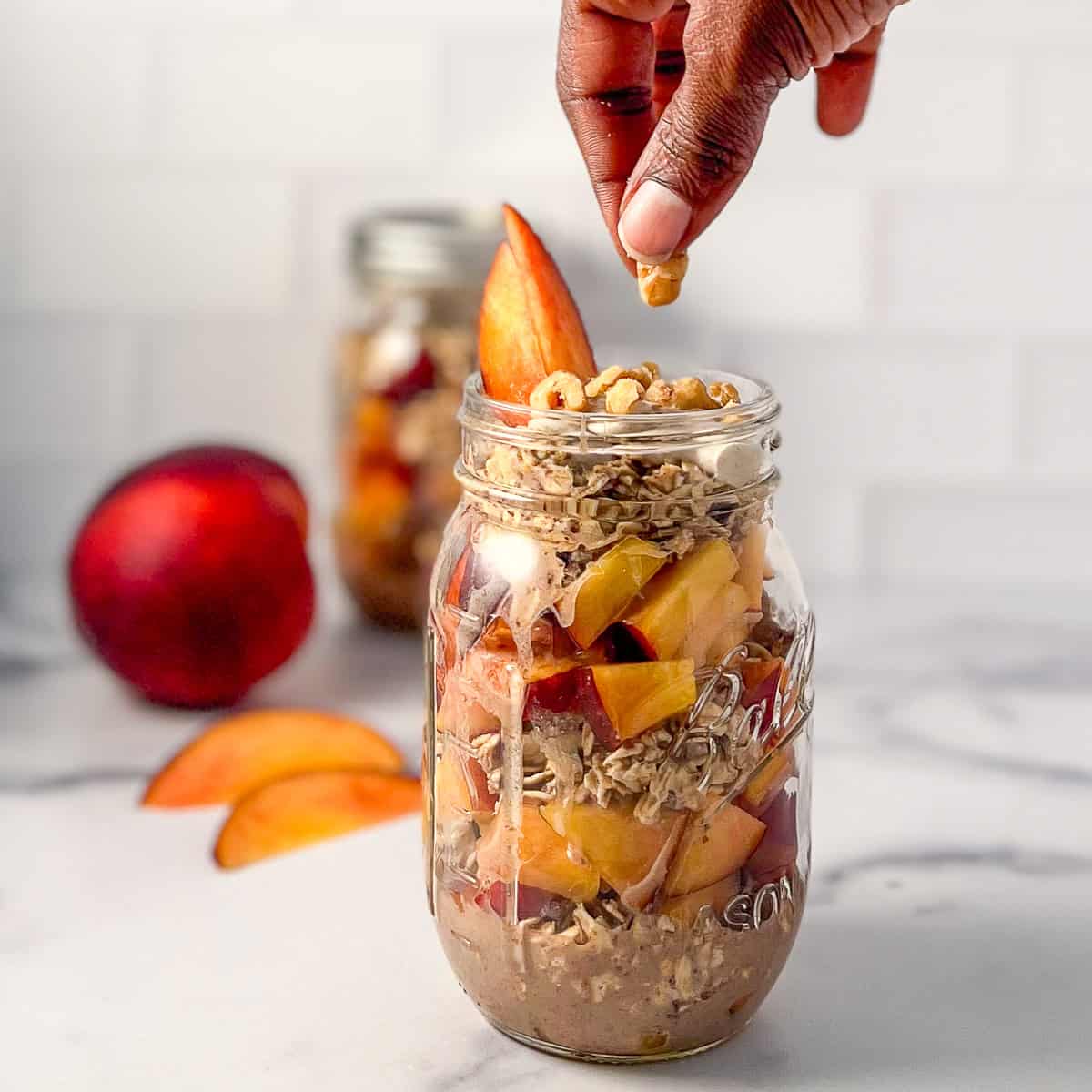 A woman's hand adding chopped walnuts to a mason jar with peach overnight oats.