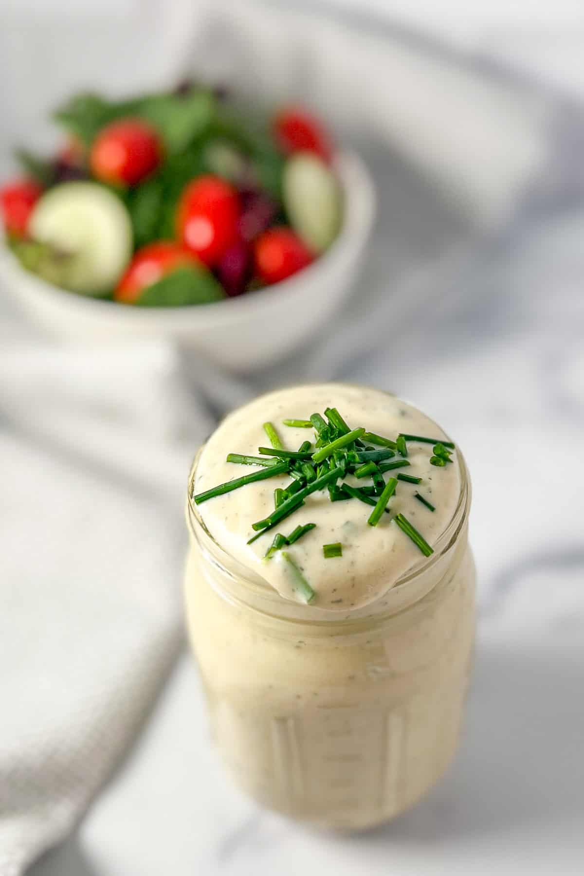 Vegan ranch dressing in a mason jar topped with fresh chopped chives and salad blurred in the background.