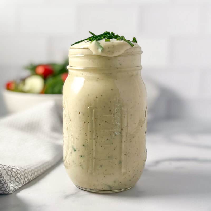Vegan ranch dressing in a mason jar topped with fresh chopped chives; salad blurred in the background.
