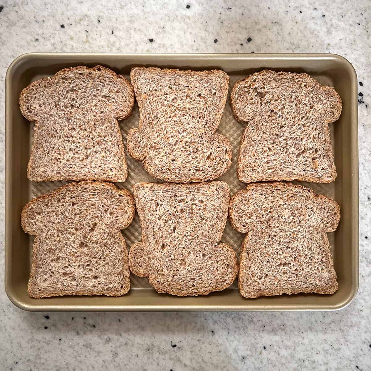 Six slices of whole grain bread arranged on a shallow baking sheet pan.