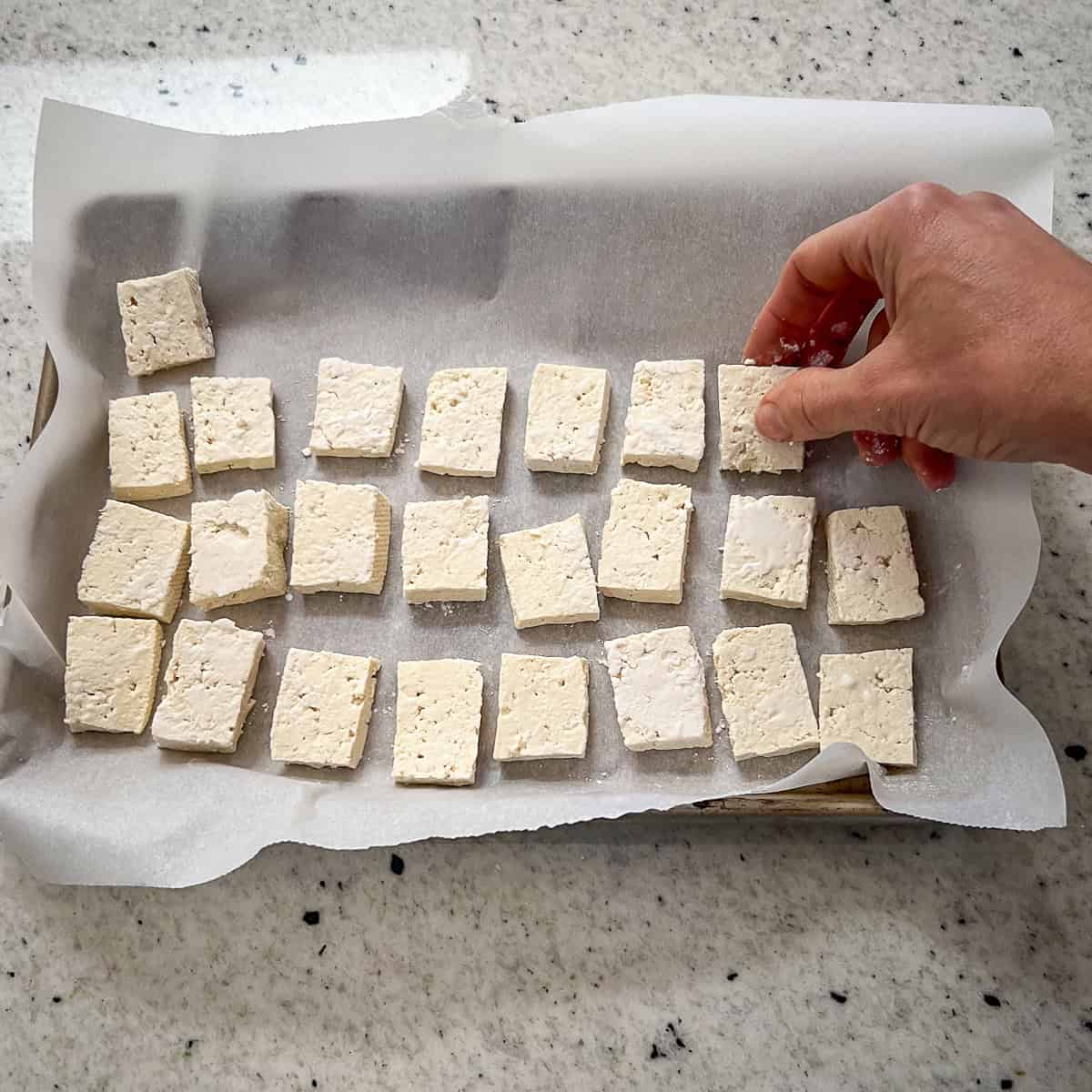 Tofu chunks being added to a parchment lined baking sheet.