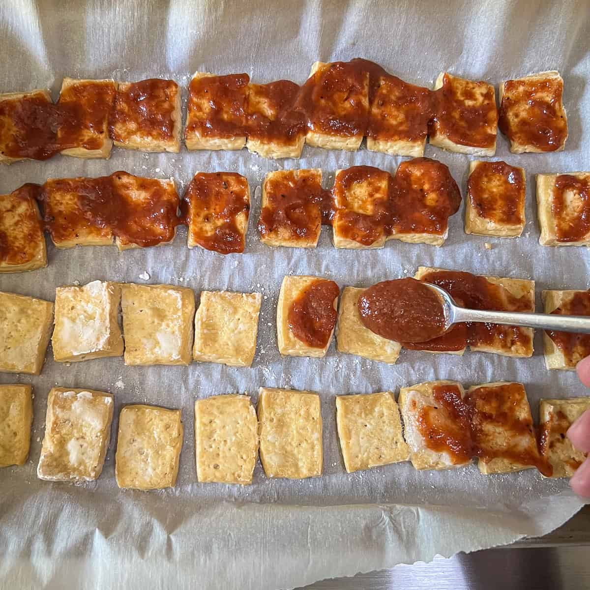 BBQ sauce being added to the baked tofu on a parchment lined baking sheet.