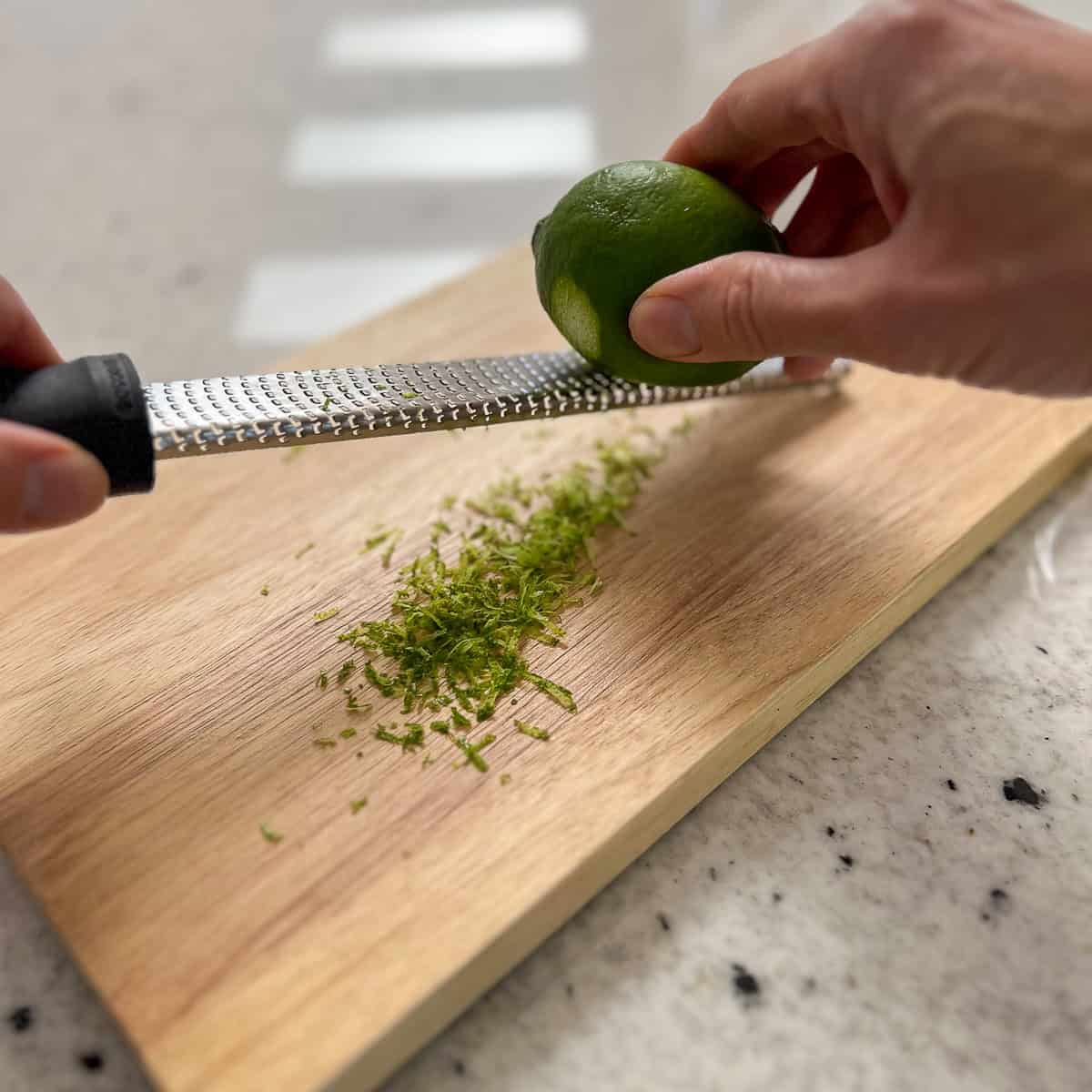 A woman's hand zesting a fresh lime on a cutting board using a microplane grater.