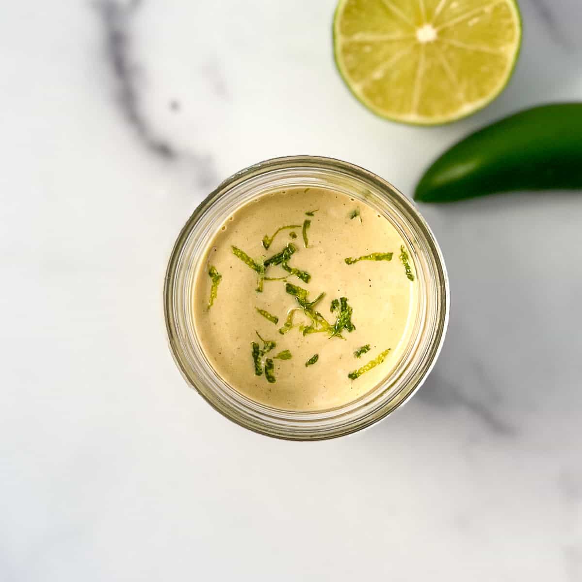 Vegan jalapeno lime aioli in a mason jar topped with fresh lime zest; lime and jalapeno blurred in the background.