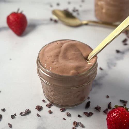 Tofu chocolate mousse in a glass dish with spoon sticking out; fresh strawberries and cacao nibs next to the dish.