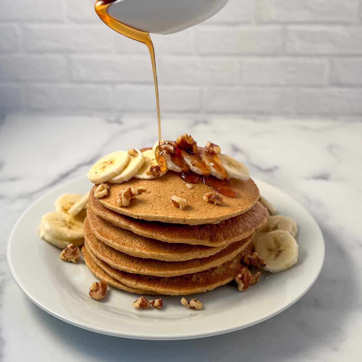 A stack of vegan banana oat pancakes with sliced banana and chopped walnuts, with maple syrup being poured over the top.