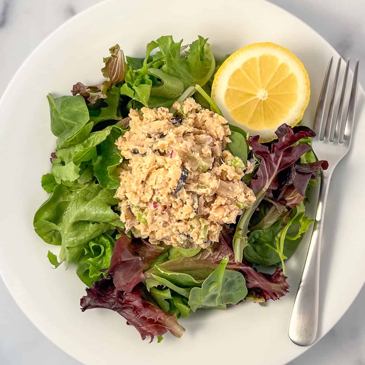 Vegan chickpea tuna salad over a bed of mixed salad greens and fresh lime wedge on the side; and fork.