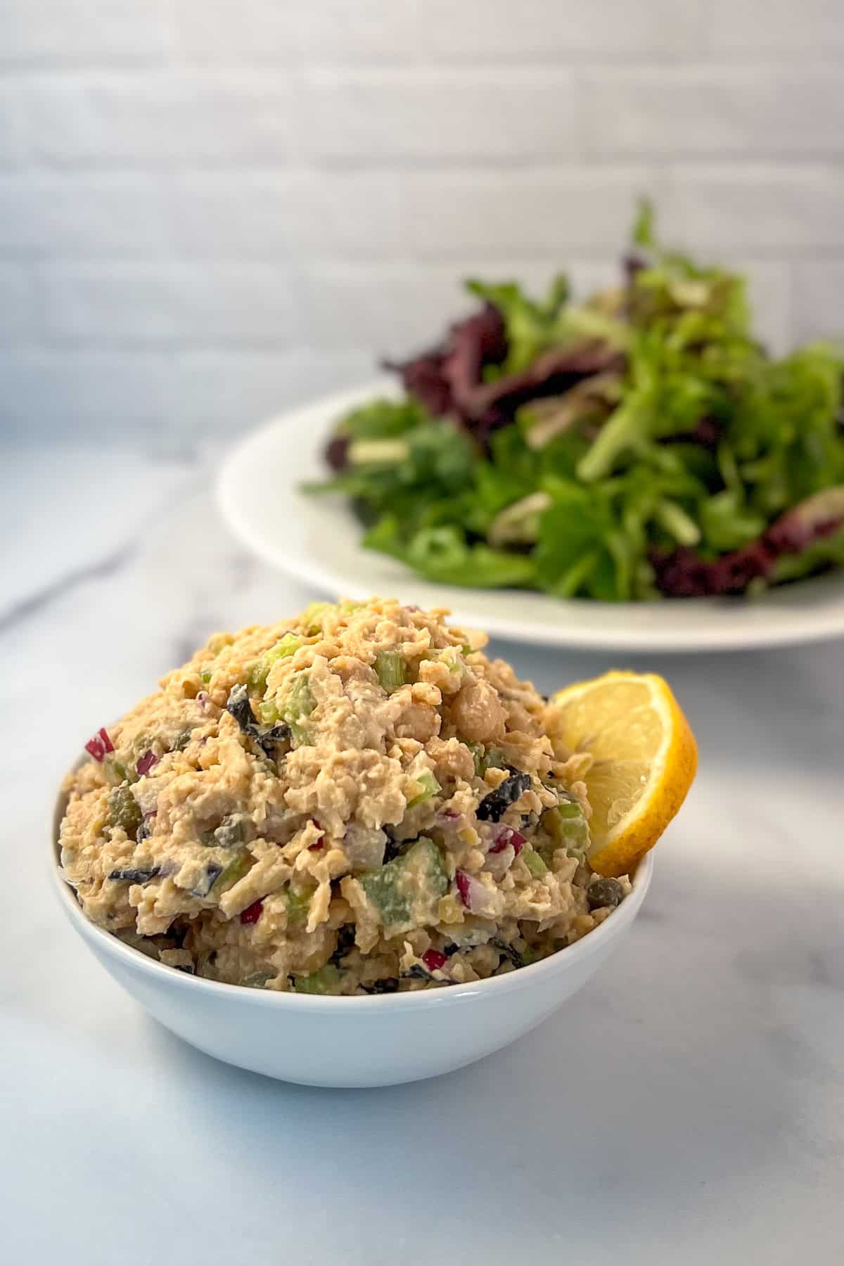 Vegan chickpea tuna salad in a bowl with lemon wedge on the side; green salad blurred in the background.