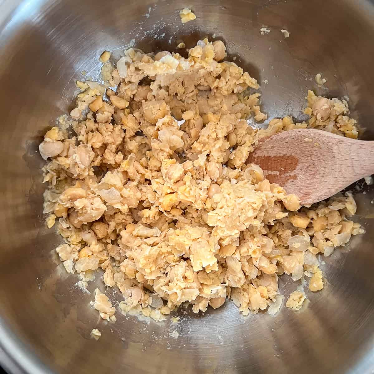 Chickpeas being smashed in a mixing bowl with a wooden spoon.