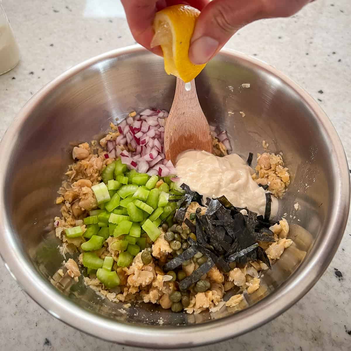 A mixing bowl with minced onion, celery, nori, vegan mayo, capers and lemon juice being added to the smashed chickpeas.