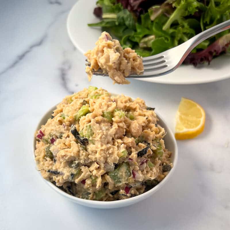 Vegan chickpea tuna salad in a bowl with a fork and salad blurred in the background.