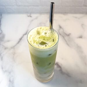 Banana milk matcha latte in a tall glass with ice and a metal straw.
