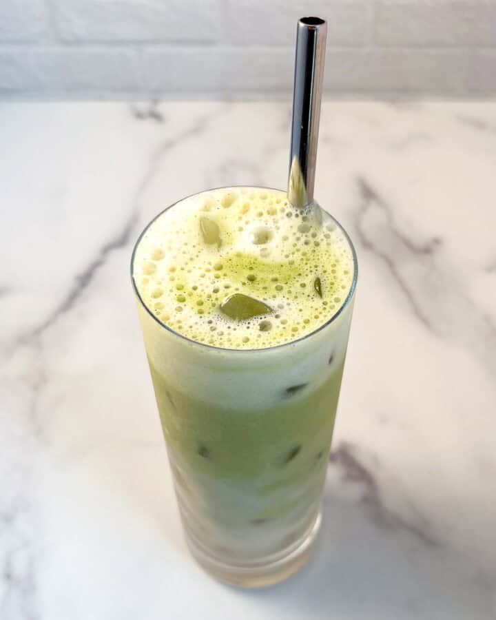 Banana milk matcha latte in a tall glass with ice and a metal straw.