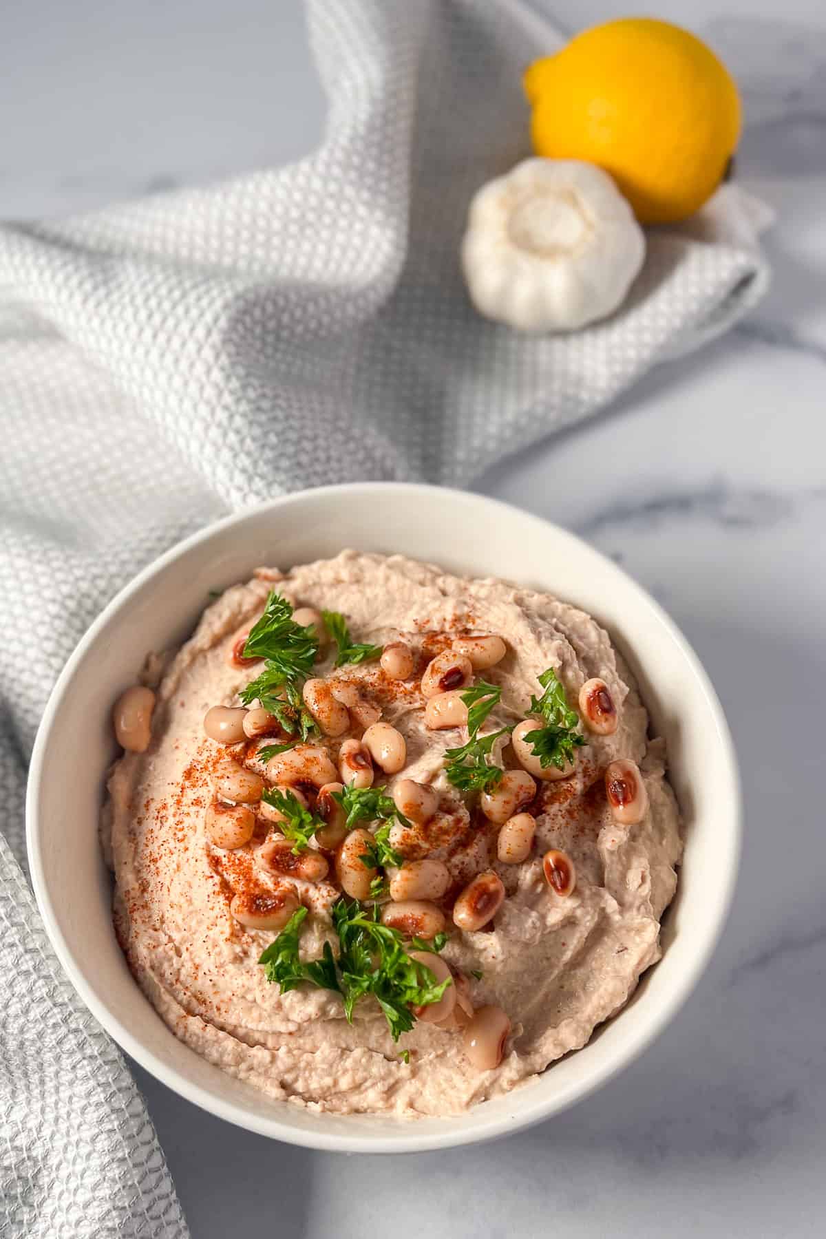 Black eyed peas hummus in a bowl topped with whole peas and fresh chopped herbs; lemon and garlic blurred in the background.