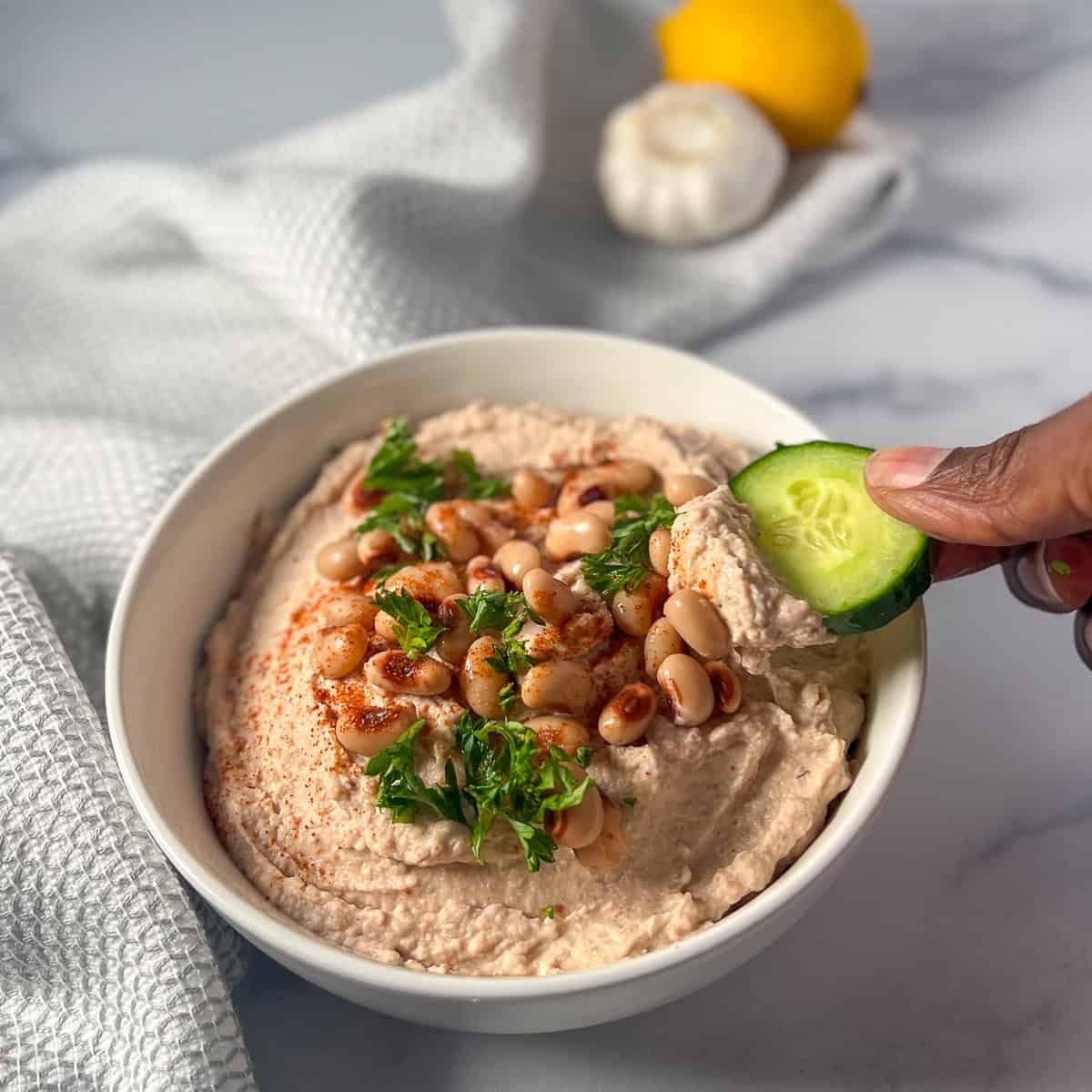 A bowl of black-eyed pea hummus with a woman's hand dipping some out with  sliced cucumber.