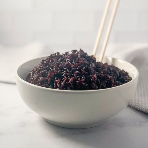 Cooked black rice in a bowl with two wooden chopsticks sticking out.