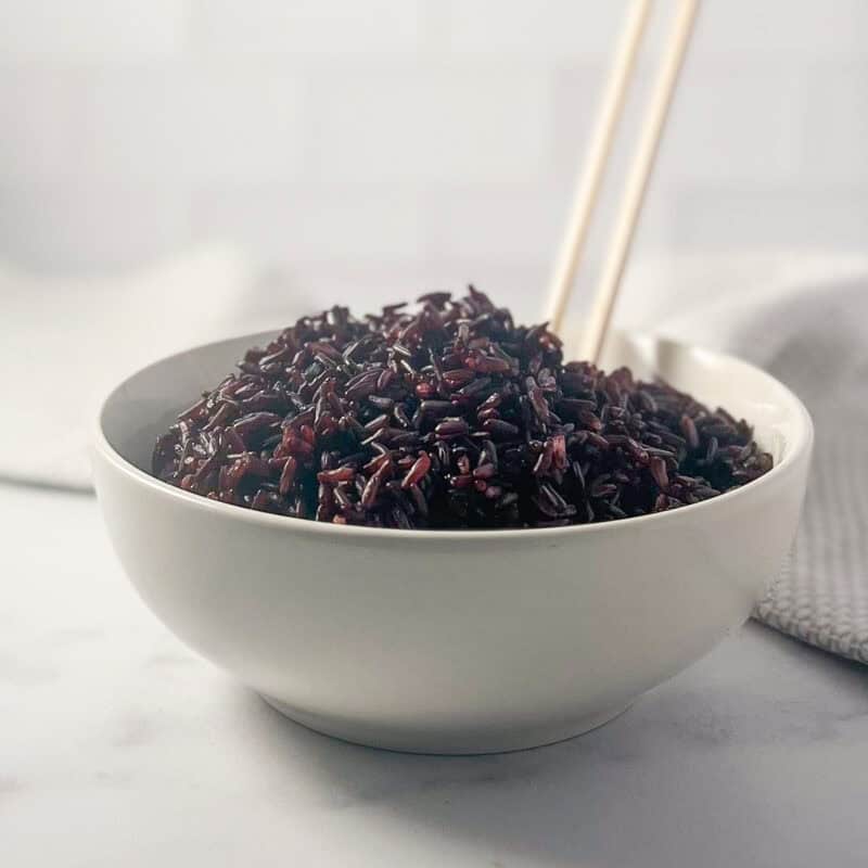 Cooked black rice in a bowl with two wooden chopsticks sticking out.