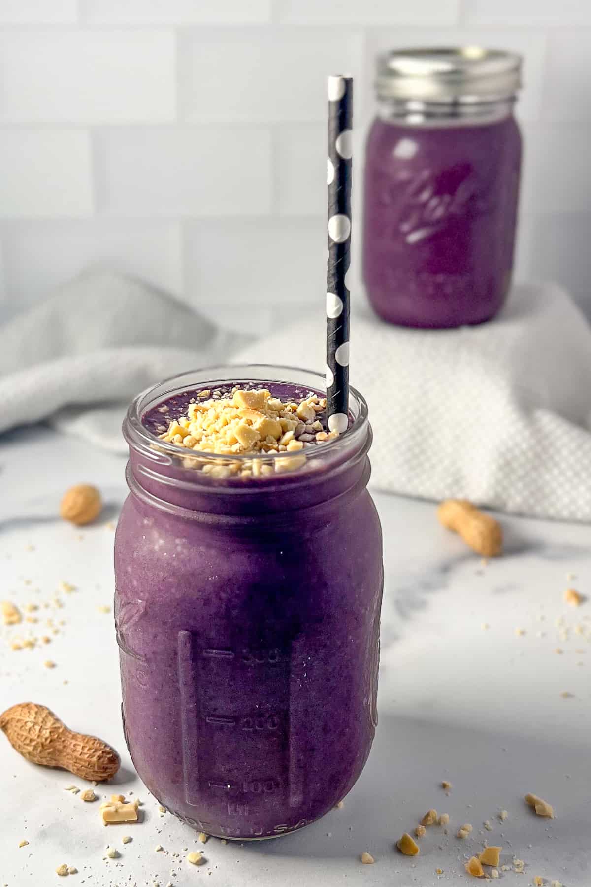 Blueberry peanut butter smoothie in a mason jar with straw topped with fresh chopped peanuts; a second smoothie blurred in the background.