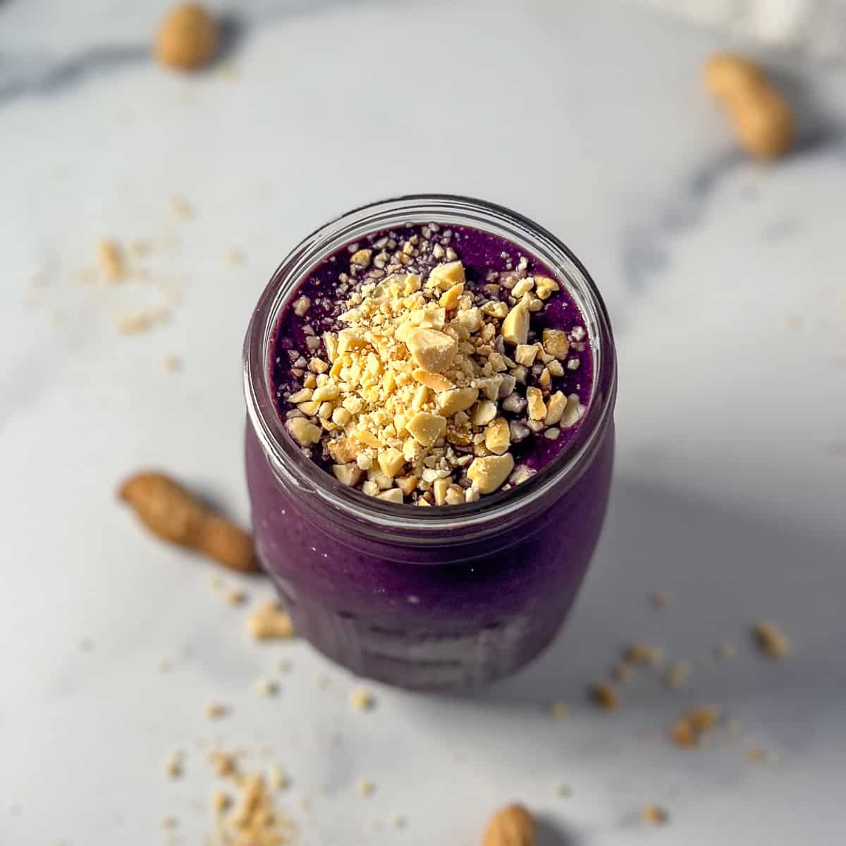 Blueberry peanut butter smoothie topped with fresh chopped peanuts; loose peanuts in the shell blurred in background.