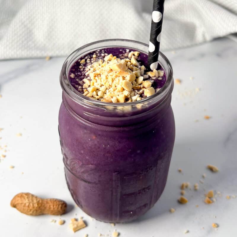 A blueberry peanut butter smoothie in a mason jar topped with chopped peanuts and a straw.