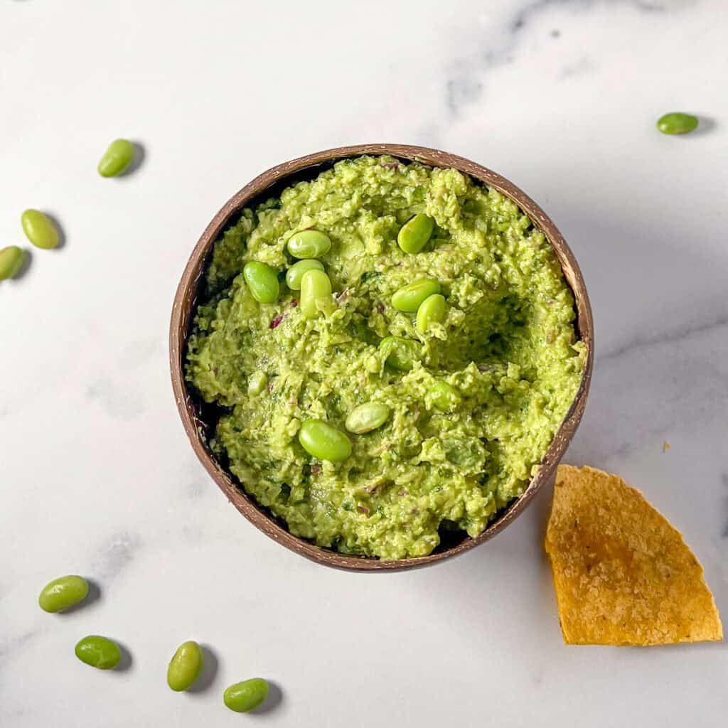A bowl of edamame hummus with a corn tortilla chip on the side.