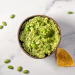 A bowl of edamame hummus with a corn tortilla chip on the side.