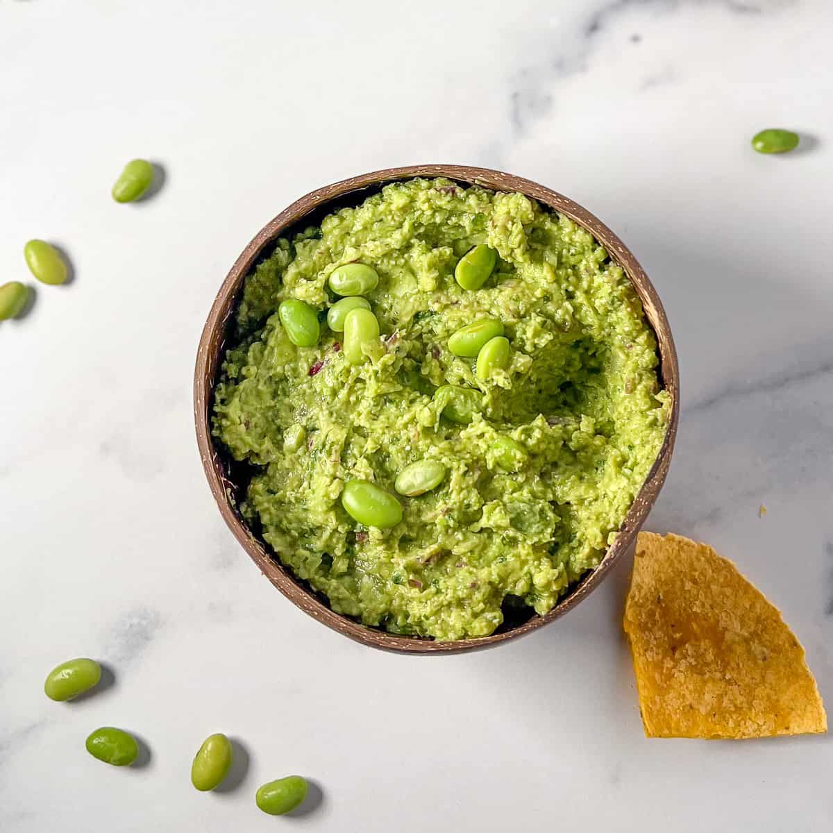 A bowl of edamame hummus with a corn tortilla chip on the side.