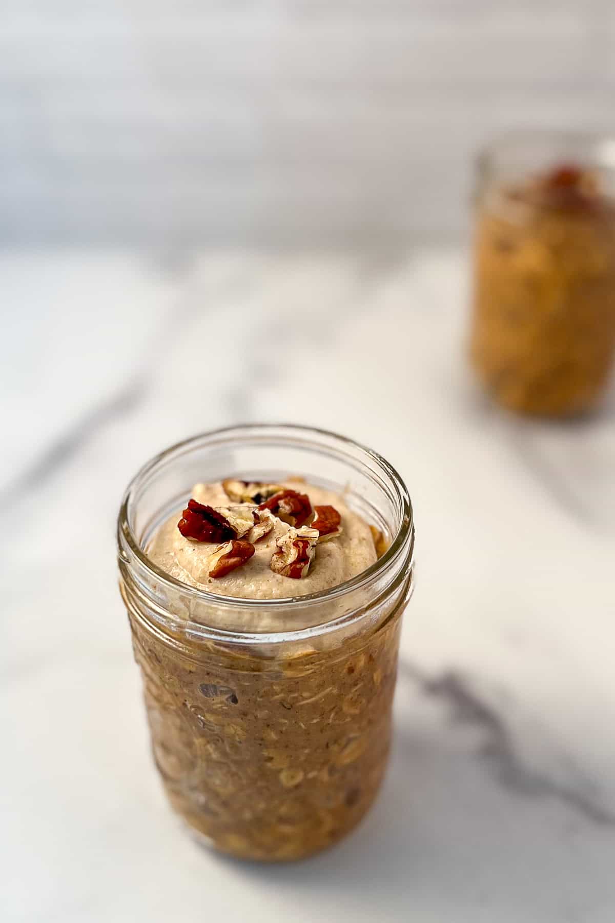 Pumpkin spice overnight oats topped with chopped pecans in a mason jar; a second jar blurred in the background.