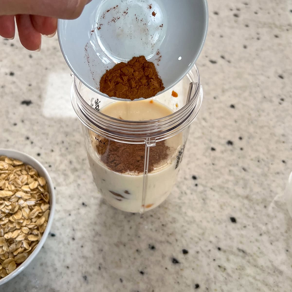 Pumpkin pie spice being added to a blender cup with plant based milk, date syrup and pumpkin puree.