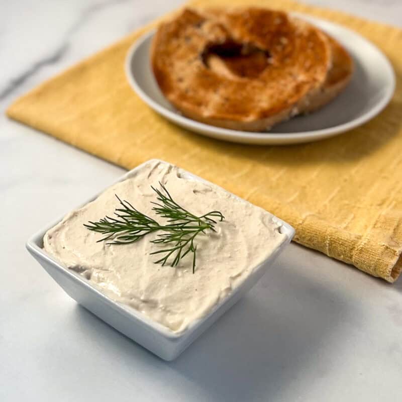 Vegan cream cheese in a square bowl with toasted bagel blurred in the background.