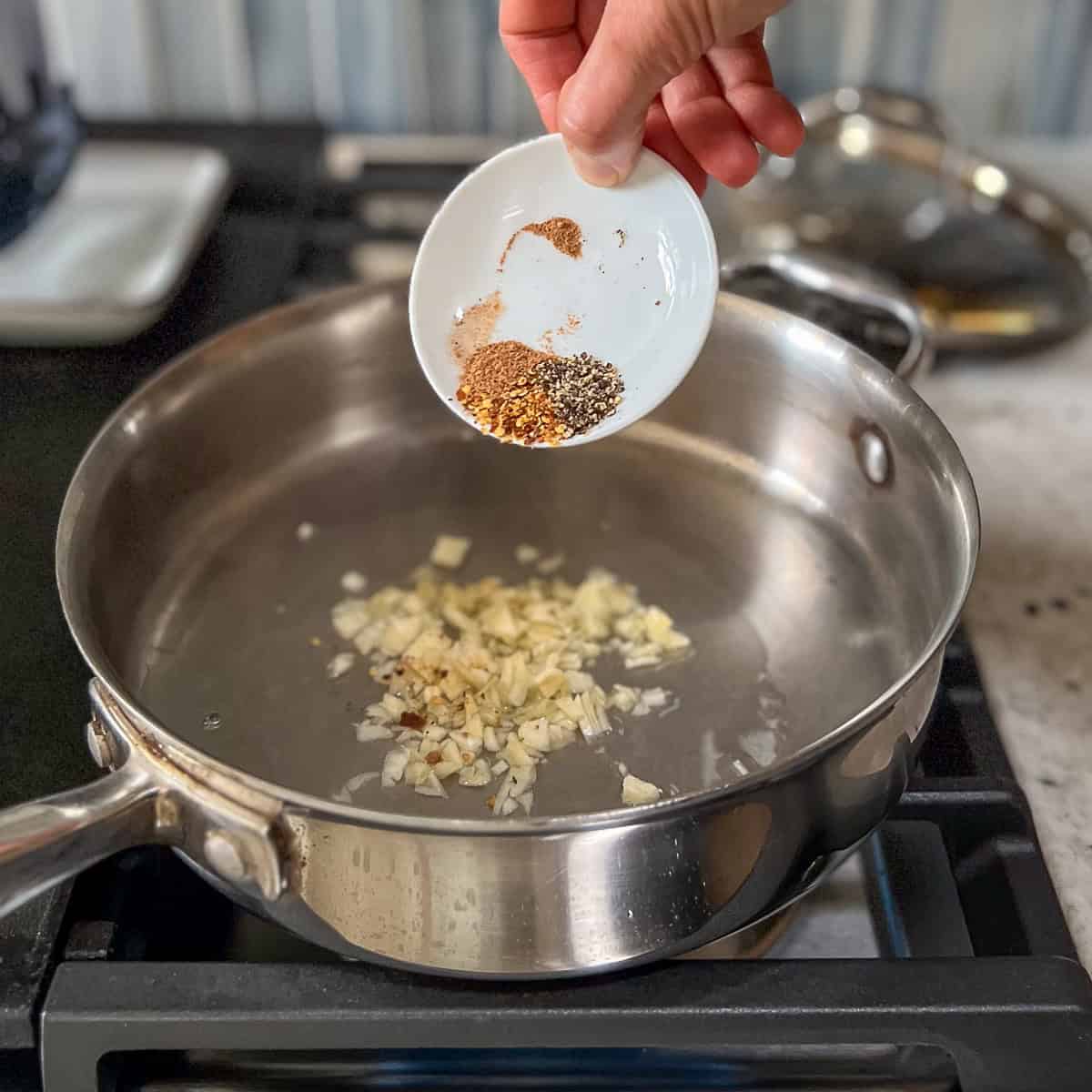 Garlic and spices being added to a hot skillet with pasta water on the stovetop.