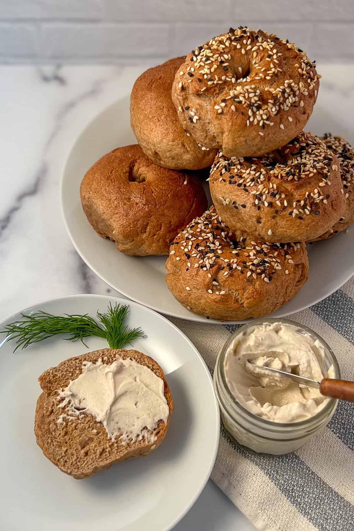 A stack of whole wheat bagels on a plate with one half on another plate with homemade cashew cream cheese.