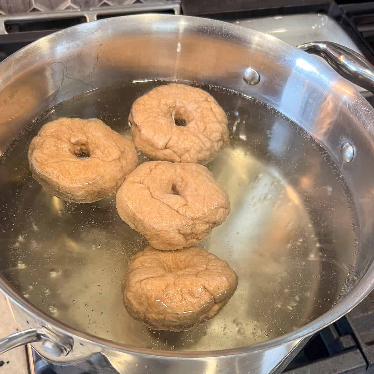 Four whole wheat bagels being boiled in water with maple syrup.