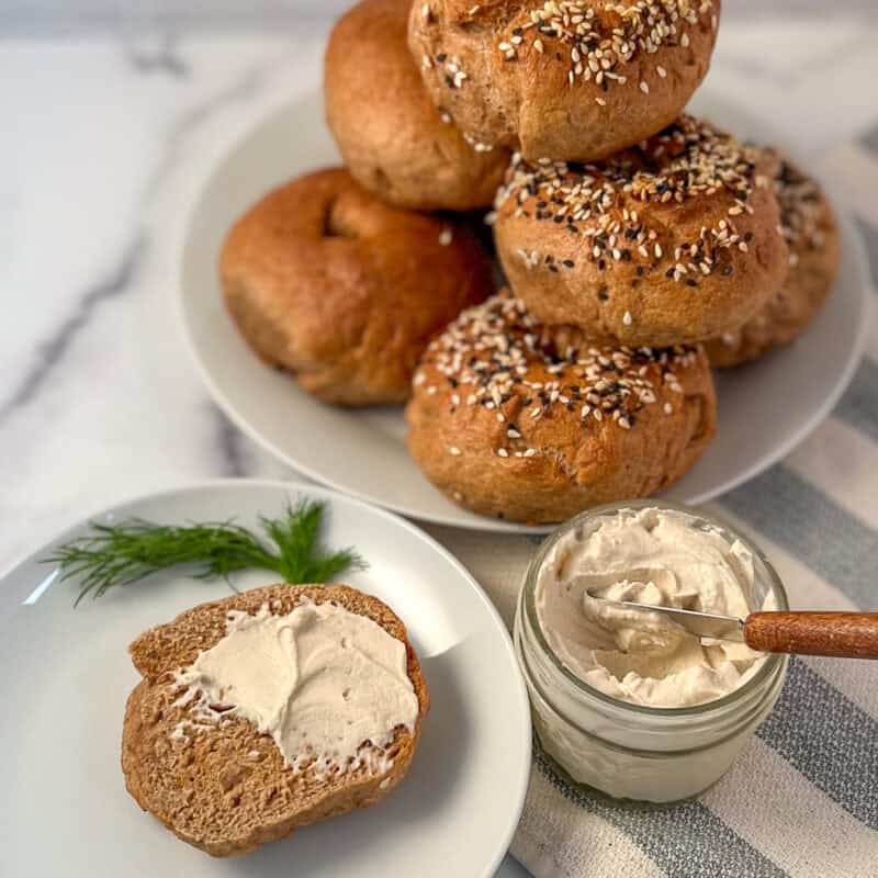 Half of a whole wheat bagel with vegan cream cheese, fresh dill and a stack of bagels on a plate in the background.