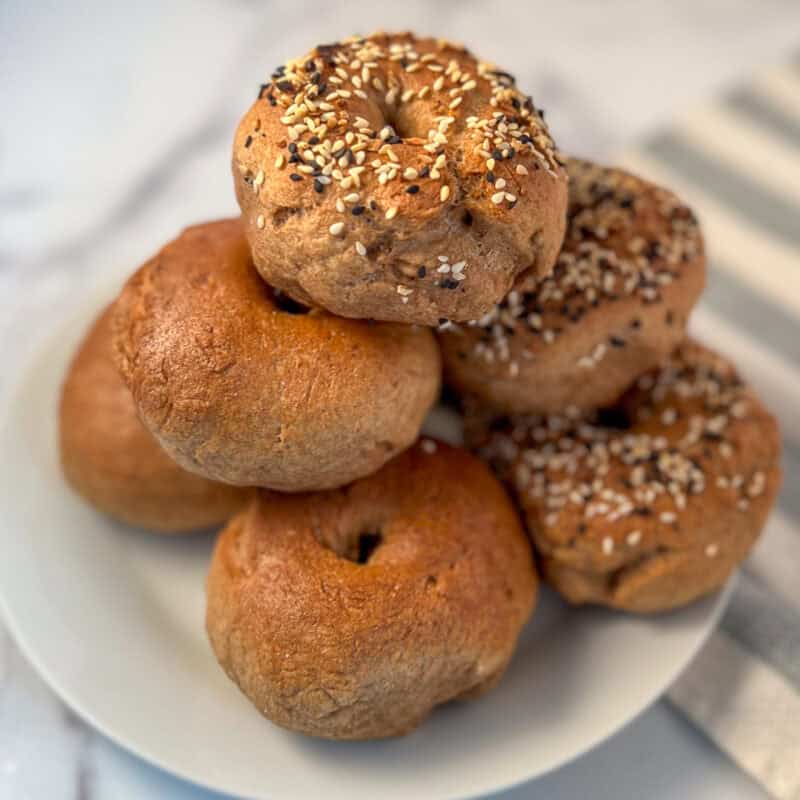 A pile of homemade whole wheat bagels on a plate, half with everything bagel seasoning on top.