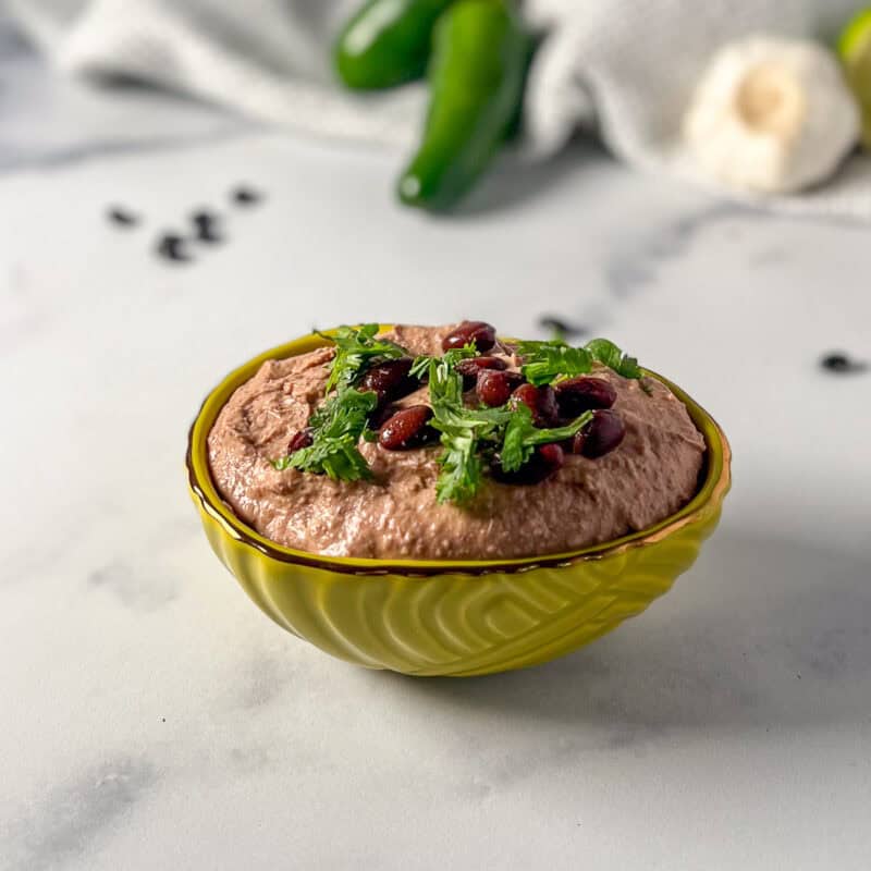 A bowl of black bean hummus topped with fresh herbs; beans, garlic and jalapeno blurred in the background.