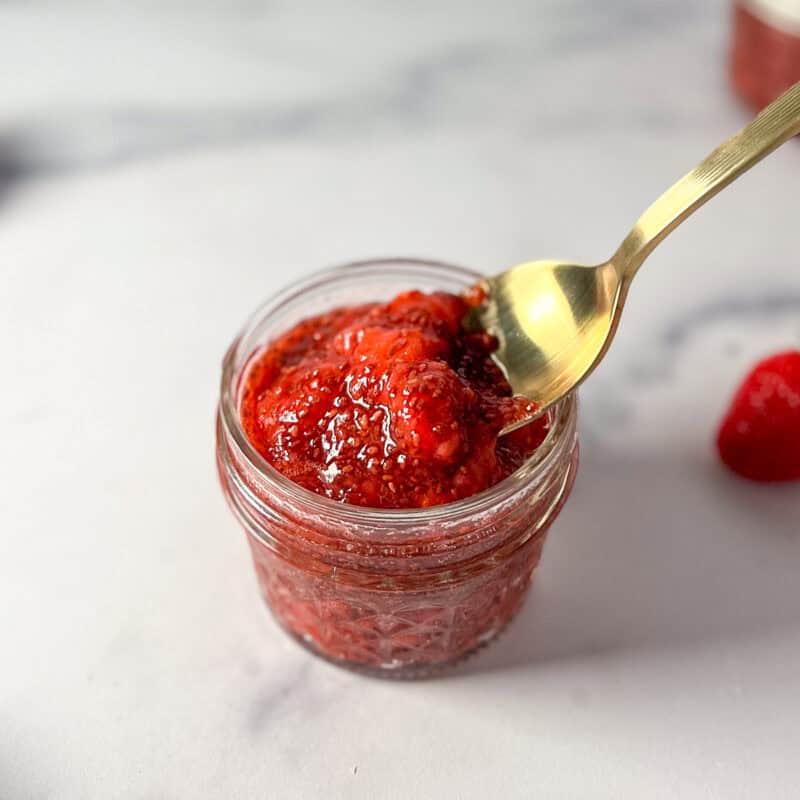 Strawberry chia seed jam in a mason jar with a spoon lifting some out to show consistency of jam.