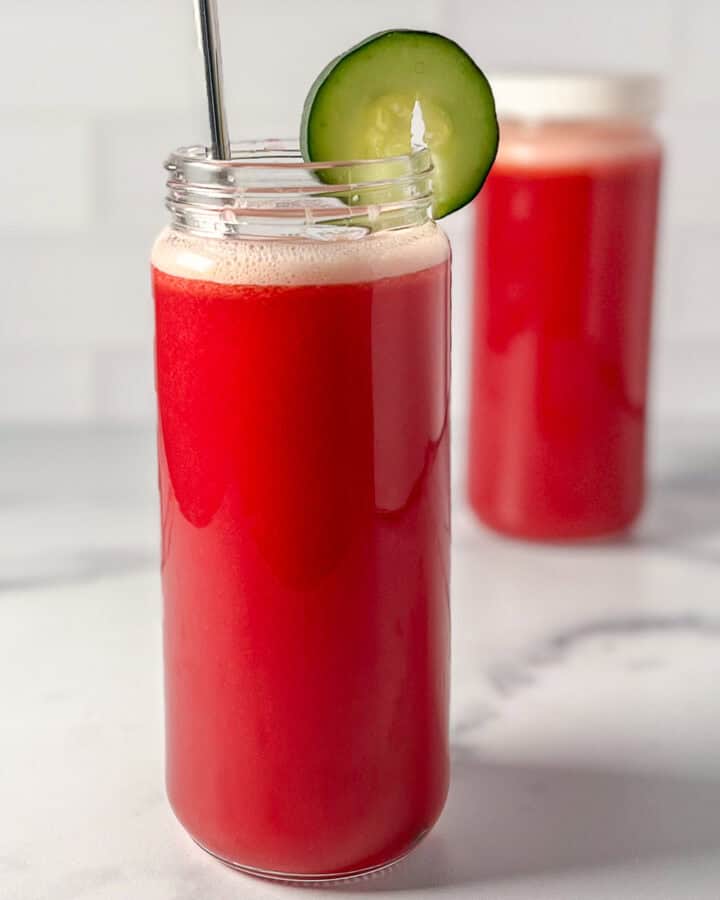 A cabbage pineapple juice in a tall glass with straw and sliced cucumber garnish.