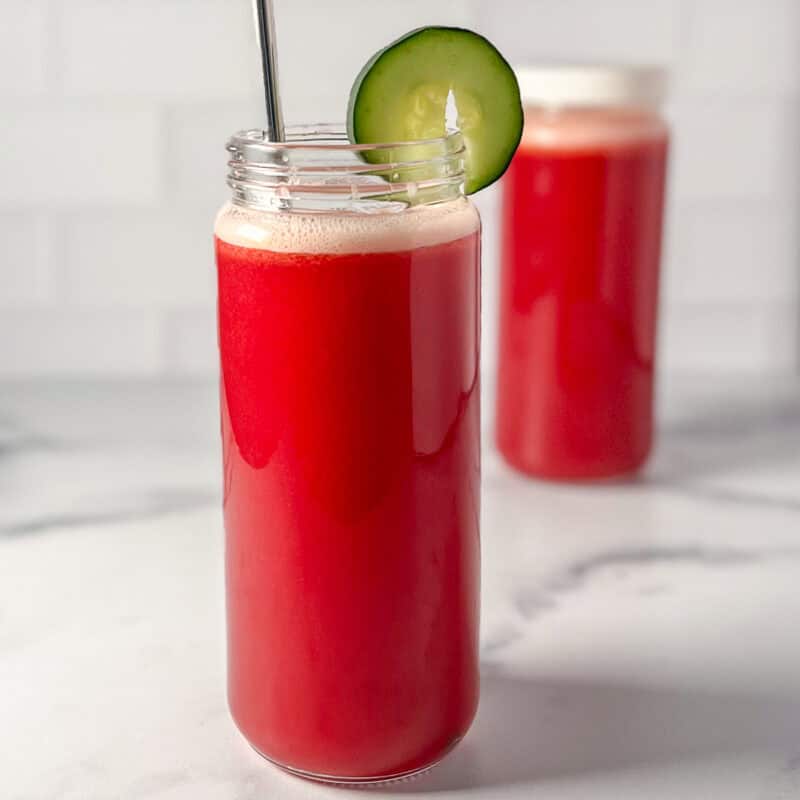 A cabbage pineapple juice in a tall glass with straw and sliced cucumber garnish.