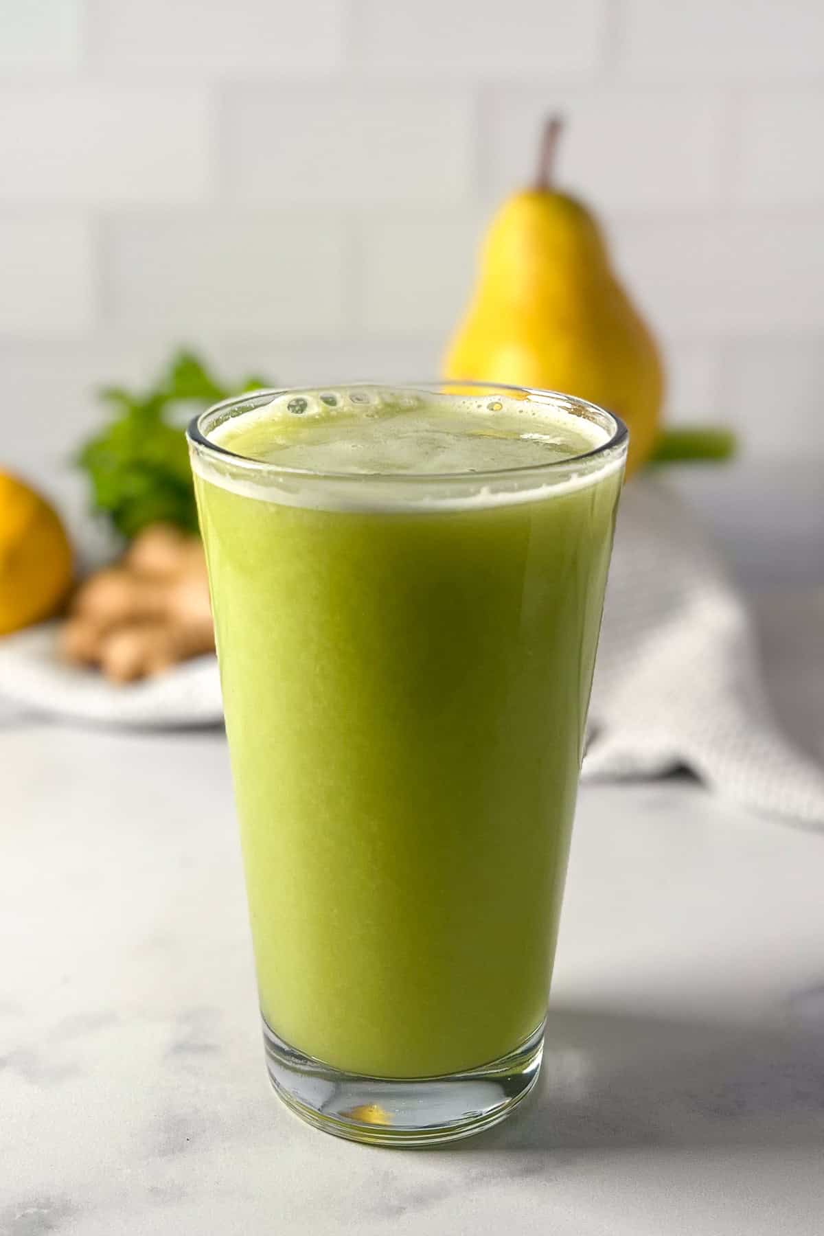 A glass of celery pear ginger juice with pear and ginger root blurred in the background.