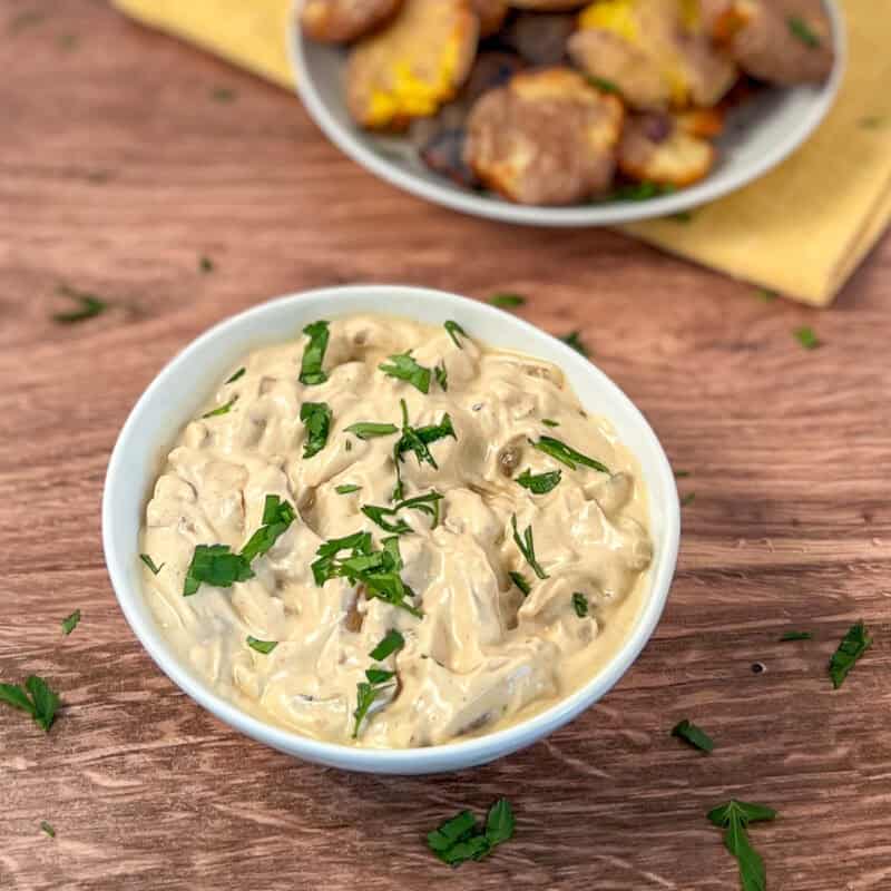 A bowl of vegan french onion dip topped with fresh chopped parsley. Air fryer smashed potatoes blurred in the background.